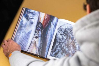 Livre de cuisine ouvert sur double page avec des image de poisson sur avec fond doré, silhouette de Pierre Manas en premier plan.